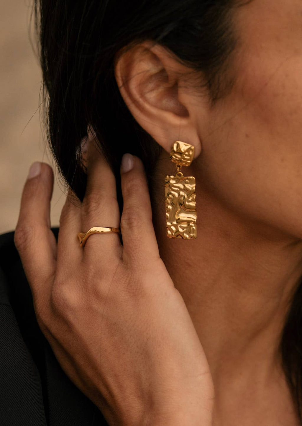 Gold hoop earrings being worn with a hand adjusting them, against a neutral background.