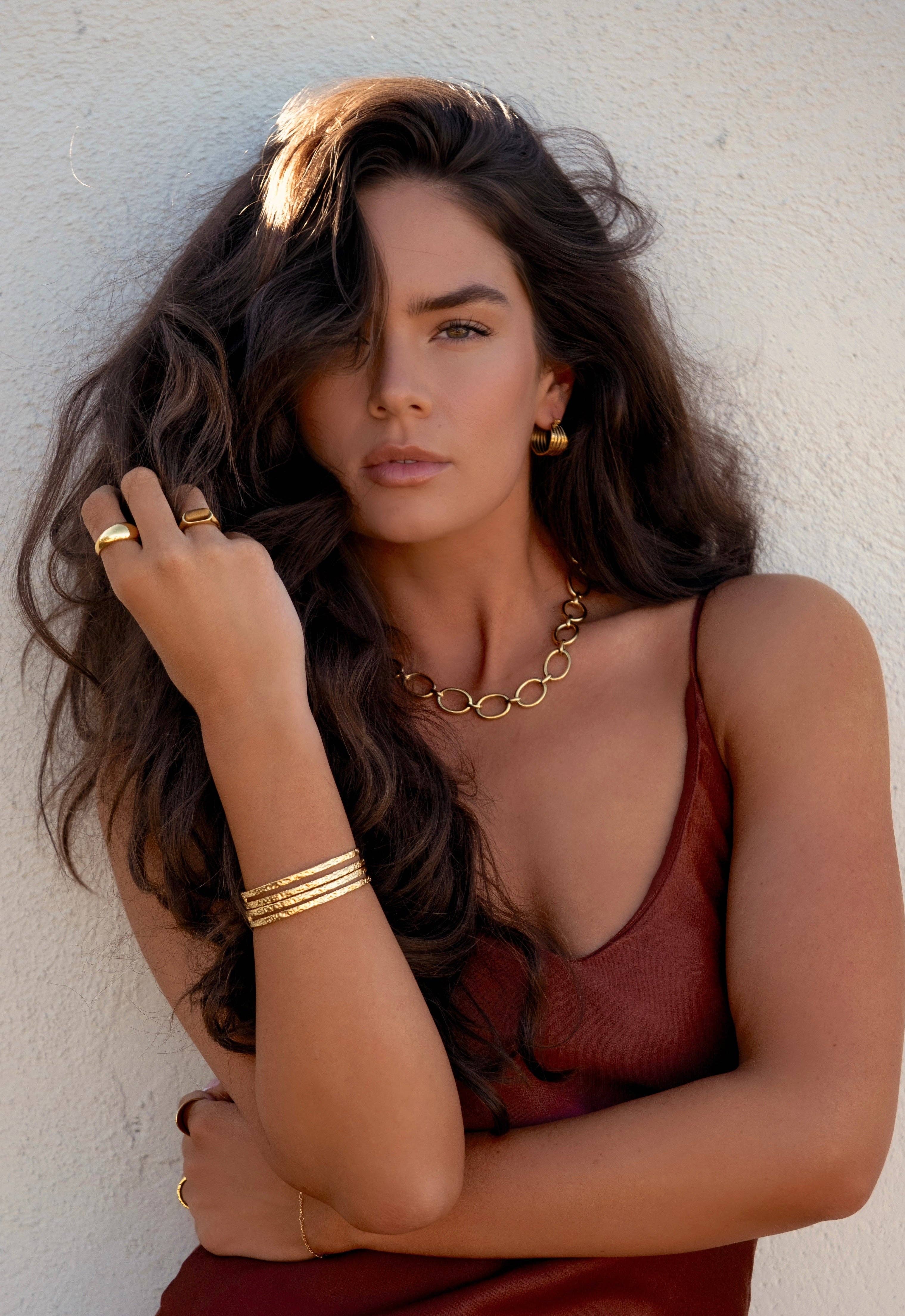 Woman with long, wavy hair wearing gold jewelry against a white wall